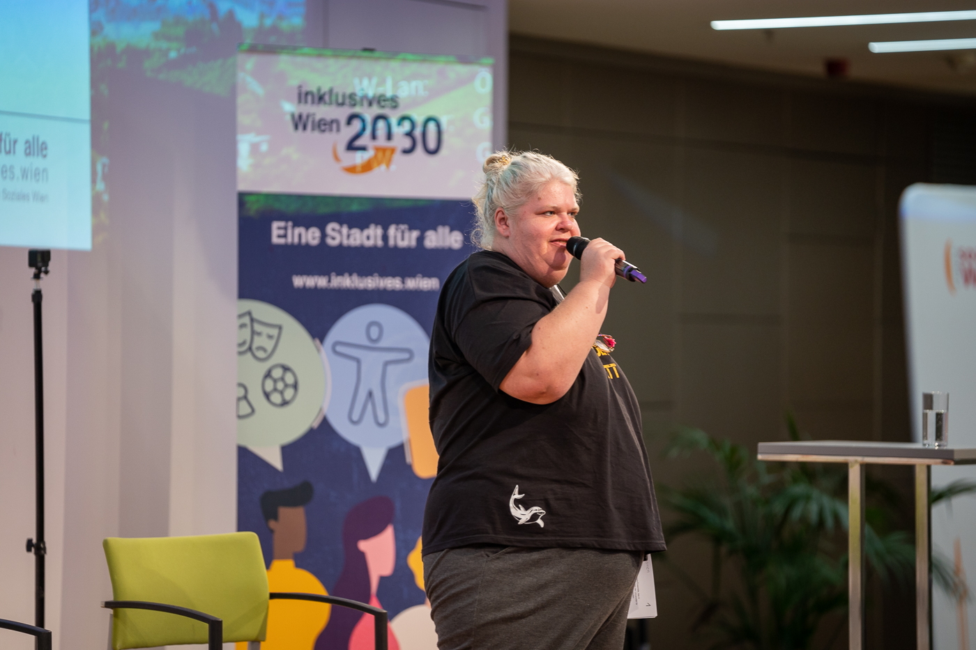 Moderatorin Iris Kopera steht vor einem Banner von "Inklusives Wien 2030" und hält ein Mikrophon in ihrer Hand.